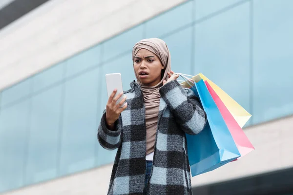 Ağzı açık tesettürlü, çantalı ve çantalı heyecanlı siyahi kadın akıllı telefondaki mesaja bakar. — Stok fotoğraf