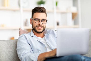 Gözlüklü yakışıklı adam evde dizüstü bilgisayar kullanıyor, koltukta oturuyor, evden çalışıyor, e-posta yazıyor ya da programlıyor.