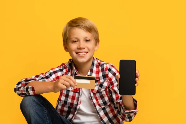Alegre guapo adolescente rubio alumno show smartphone con pantalla en blanco y tarjeta de crédito — Foto de Stock