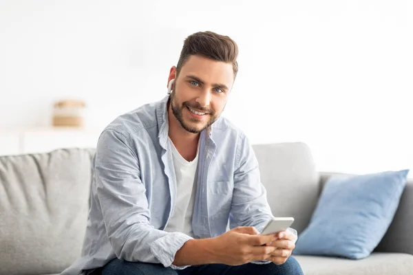 Jóképű Y-generációs férfi fülhallgatóban okostelefonnal és mosollyal a kamerába, otthon ül a kanapén, másolja a helyet — Stock Fotó
