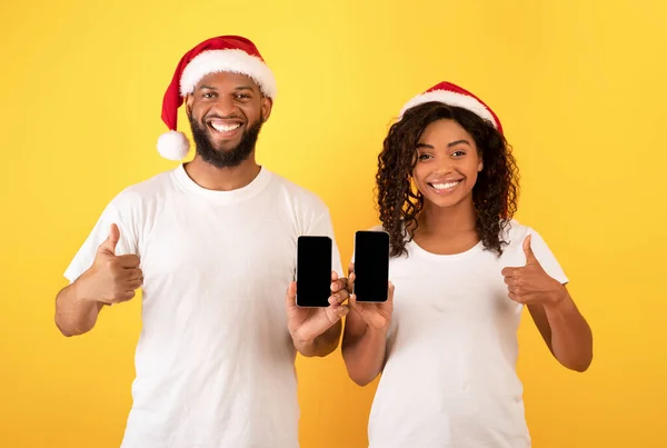 Mutlu Afro-Amerikalı çift ellerinde boş ekranlı akıllı telefonlar tutuyorlar ve başparmaklarını gösteriyorlar, sarı arka plan. — Stok fotoğraf