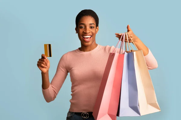 Alegre hembra negra con bolsas de compras y tarjeta de crédito sobre fondo azul —  Fotos de Stock