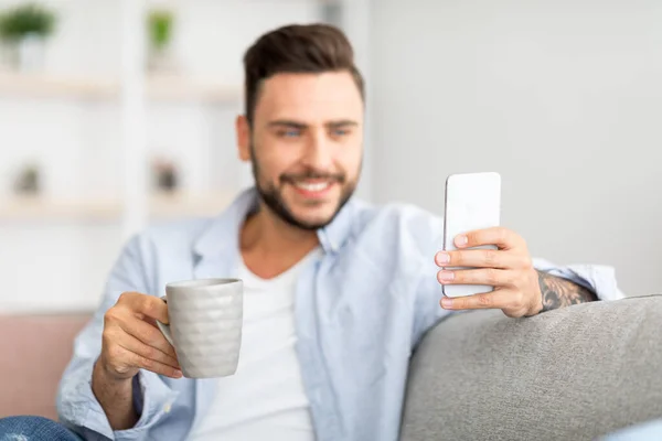 Concepto de vida en línea. Emocionado hombre milenario sosteniendo teléfono inteligente moderno y taza de café en casa, espacio libre — Foto de Stock