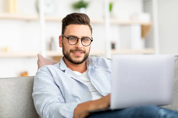 Bonito cara com óculos usando laptop em casa, sentado no sofá, trabalhando em casa, digitando e-mails ou programação — Fotografia de Stock