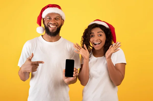 Julförsäljning online. Spännande afrikansk amerikansk par pekar på tom smartphone skärm över gul bakgrund — Stockfoto