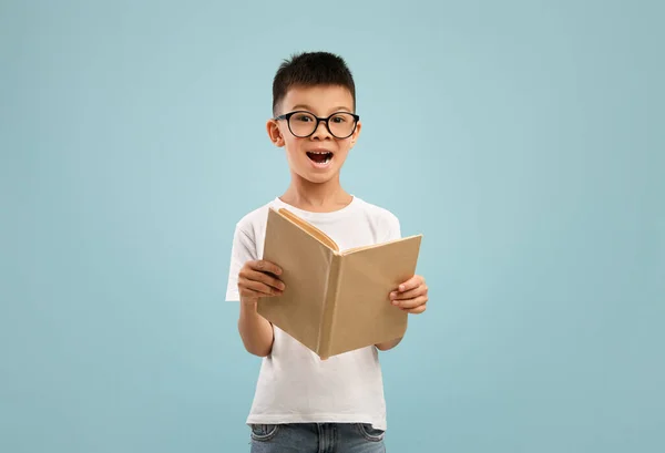 Aufgeregt kleiner asiatischer Junge mit Brille hält und liest aufgeschlagenes Buch — Stockfoto