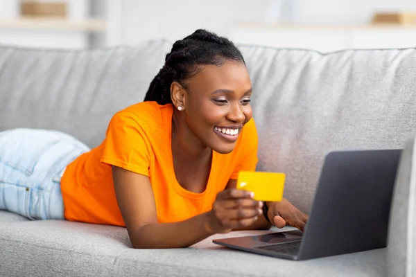 Neşeli Afro kadın internetten kredi kartı ve laptopuyla alışveriş yapıyor, evdeki kanepede yatıyor. — Stok fotoğraf