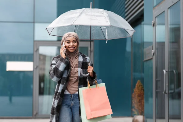 Щаслива африканська жінка в Хіджабі і пальто з пакетами закупівель і парасольки біля магазину. — стокове фото