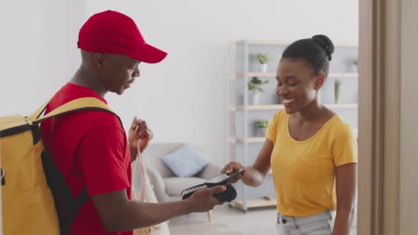 Cheerful african american woman paying for food order from market with smartphone, getting bag from courier at home — Stock Video