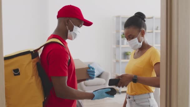 Koruyucu maskeli genç Afro-Amerikan bayan akıllı telefonla ödeme yapıyor ve kuryeden sipariş paketi alıyor. — Stok video