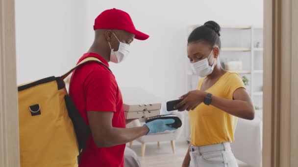 Mensajero afroamericano recibiendo pago y dando orden de pizza a cliente femenino en casa, usando máscaras protectoras — Vídeos de Stock