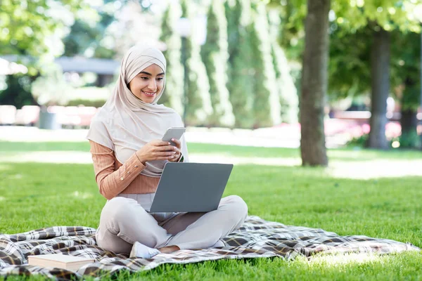 Feliz bastante joven islámica hembra en hijab con ordenador portátil charlando por teléfono, trabajando en el parque — Foto de Stock