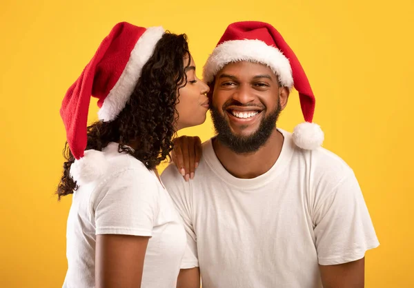 Aimer femme afro-américaine dans Santa chapeau embrasser son mari souriant, posant sur fond de studio jaune — Photo