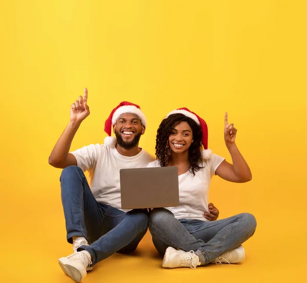 クリスマスオファー。幸せなアフリカ系アメリカ人のカップルがノートパソコンとフリースペース、黄色の背景を指して座って — ストック写真