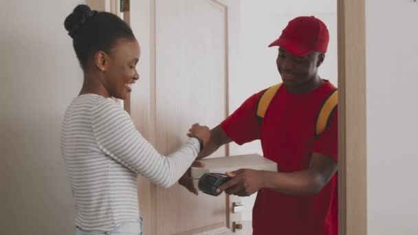 Sorridente ragazzo corriere nero con indosso uniforme rossa contenente scatole di pizza, donna afro-americana che paga con smartphone — Video Stock