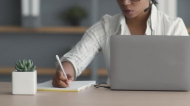 Tanımlanamayan kadın ofis yöneticisi dizüstü bilgisayarlı görüntülü sohbet yoluyla müşteriyle konuşurken not alıyor, iş yerinde oturuyor