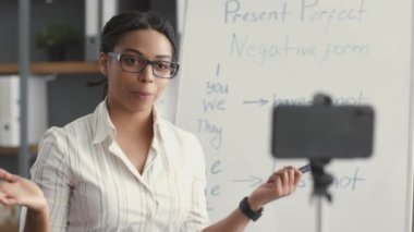 Genç, neşeli, Afro-Amerikan kadın öğretmen akıllı telefondan video kaydediyor, sınıfta poz veriyor.
