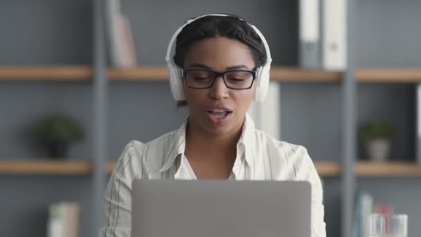 Genç duygusal Afrikalı Amerikalı bayan kulaklıkla müzik dinliyor, iş sırasında laptopta şarkı söylüyor ve dans ediyor. — Stok video