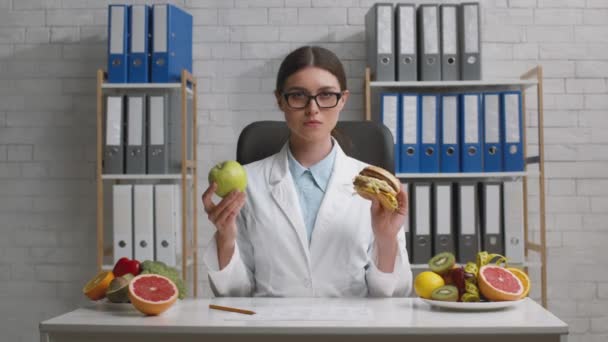 Professionele dame voedingsdeskundige tonen groene appel en junk burger om camera, het vergelijken van gezonde en ongezonde voeding — Stockvideo