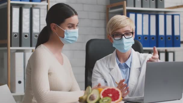 Médico nutricionista maduro y paciente con máscaras protectoras sentado en la oficina y discutiendo plan de dieta en el ordenador portátil — Vídeo de stock