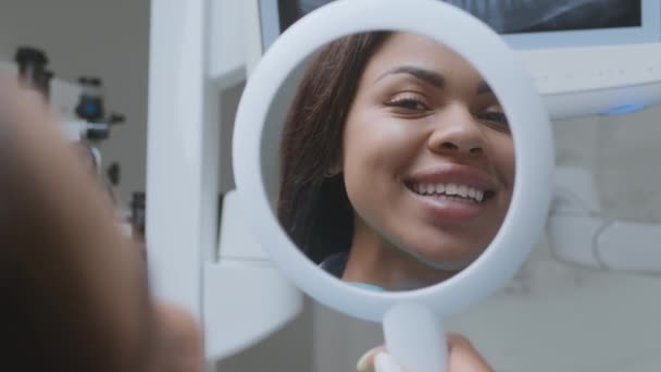 Młoda, pogodna, afrykańska kobieta patrząca w lustro i ciesząca się dobrym rezultatem wyrównywania zębów w klinice stomatologicznej — Wideo stockowe