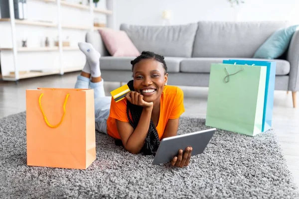 Позитивна чорна жінка лежить на підлозі з планшетом і кредитною карткою, покупки в Інтернеті з дому — стокове фото