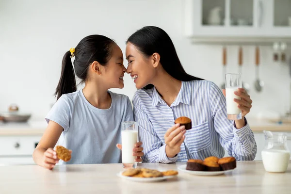 Asyalı anne ve küçük kızı sevmek mutfakta abur cubur yiyip süt içmek, yeni pişmiş keklerin tadını çıkarmak. — Stok fotoğraf