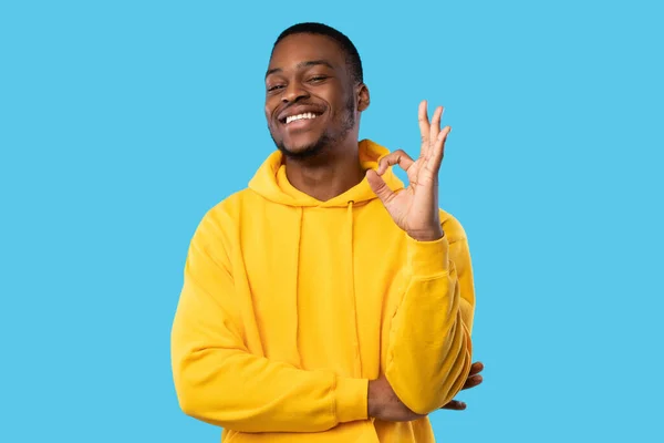 Homem negro alegre em amarelo Hoodie Gesturing Ok, fundo azul — Fotografia de Stock