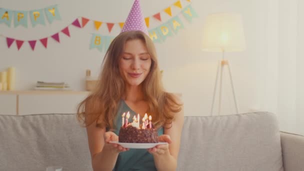 Молода весела жінка в вечірній кепці, яка дме свічки на торт, насолоджуючись святкуванням вдома, відстежуючи постріл — стокове відео