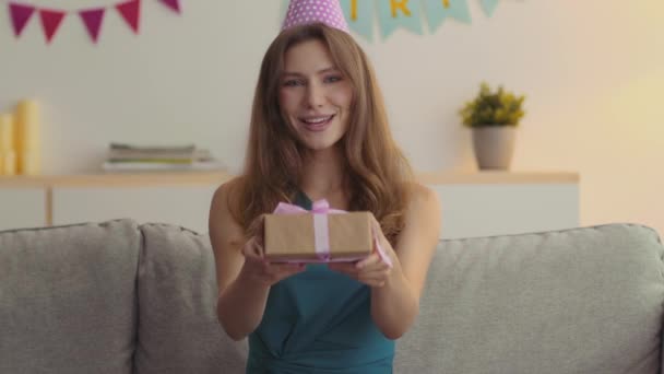 Schattige mooie jonge dame in paty cap het aanbieden van geschenkdoos om camera en zeggen Gelukkige verjaardag, zittend in versierd huis — Stockvideo