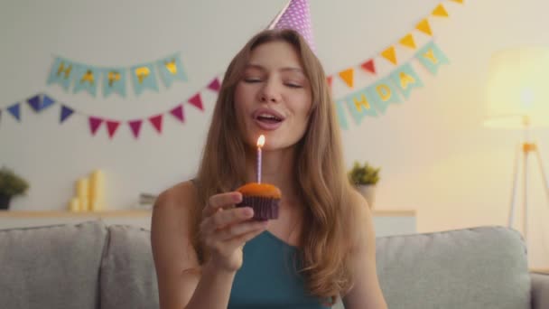 Fiesta de cumpleaños en línea. Webcam retrato de feliz joven mujer usando partido tapa soplando vela en cupcake y bailando — Vídeo de stock