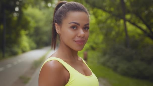 Ritratto ravvicinato di giovane donna afroamericana sportiva positiva che sorride alla telecamera, posa nel parco pubblico, inseguimento delle riprese — Video Stock