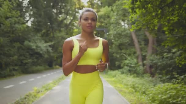 Giovane donna afroamericana sportiva in tuta sportiva brillante in esecuzione al mattino, guardando la fotocamera, facendo jogging nel parco pubblico — Video Stock