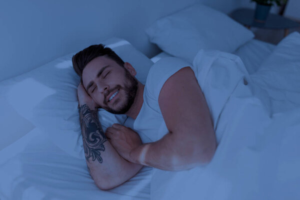 Happy millennial man lying on white linen flax bedsheets and smiling, relaxing in bedroom at night, free space