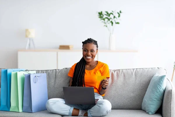 Hermosa señora Afro compras en línea, sentado en el sofá con la PC portátil, la celebración de la tarjeta de crédito, bolsas de regalo de pie cerca — Foto de Stock