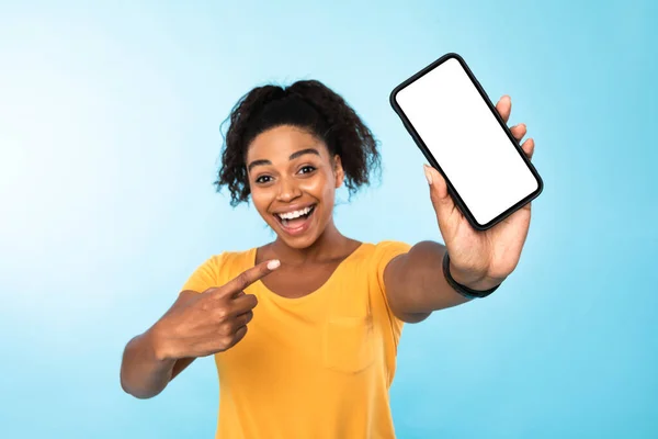 Heyecanlı siyah kadın mavi arka planda boş ekranlı akıllı telefonu işaret ediyor. Mobil uygulama, web sitesi için model — Stok fotoğraf