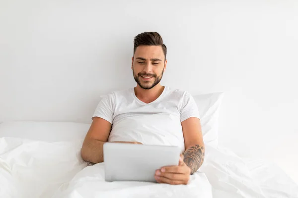 Portret szczęśliwego mężczyzny siedzącego na łóżku pod kocem i korzystającego z komputera, wpisującego na klawiaturze laptopa, wolna przestrzeń — Zdjęcie stockowe