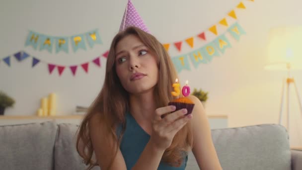 Печальный процесс старения. Молодая расстроенная женщина в кепке задувает 30 свечей на кексе, чувствуя стресс из-за своего возраста — стоковое видео