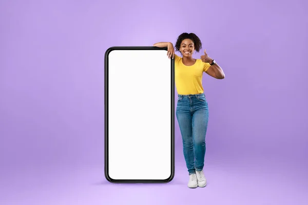 Modern akıllı telefonun yanında duran siyah kadın boş ekranla başparmağını menekşe arka planında gösteriyor. — Stok fotoğraf