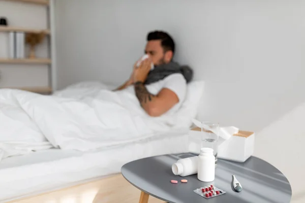 Nemocný tisíciletý muž kýchající v papírové tkáni, ležící pod přikrývkou v posteli. Zaměřte se na noční stolek plný léků — Stock fotografie