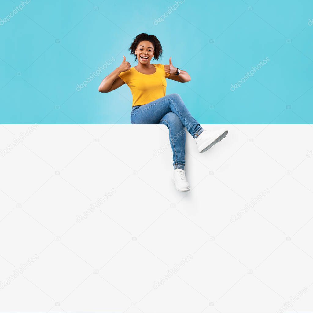 Excited young black lady sitting on blank white paper banner with mockup for ad, showing thumb up on blue background