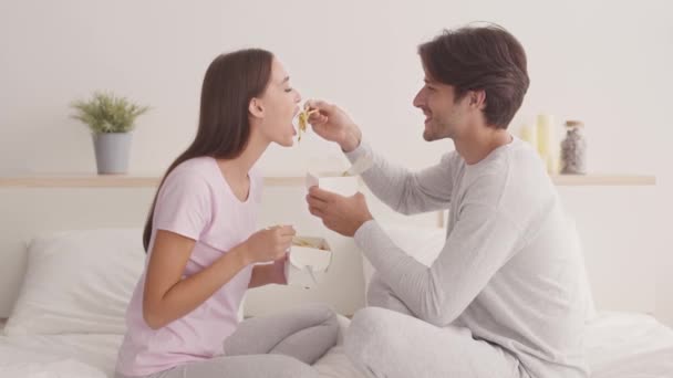 Unga lyckliga gifta par äter asiatisk mat i sängen, mannen matar sin flickvän, makar njuter av mat leverans service — Stockvideo