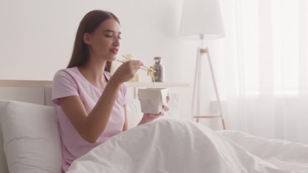 Dostawa żywności. Młoda pozytywna kobieta jedząca chiński makaron z pudełka pałeczkami, siedząca rano w łóżku — Wideo stockowe