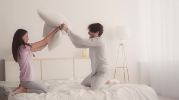 Hushållsromantik. Ung lycklig man och kvinna som har roligt hemma, leker kuddkrig och skrattar, sitter i sängen — Stockvideo