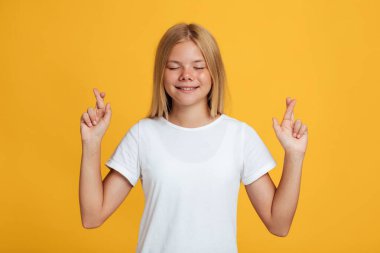 Happy cute adolescent caucasian blonde girl with closed eyes raising crossed fingers and make wish clipart