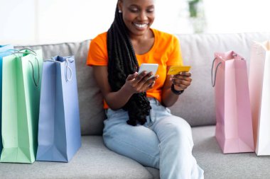 Kredi kartı ve cep telefonu olan neşeli Afro kadın kanepeye oturmuş hediye çantalarıyla online alışveriş uygulaması kullanıyor.