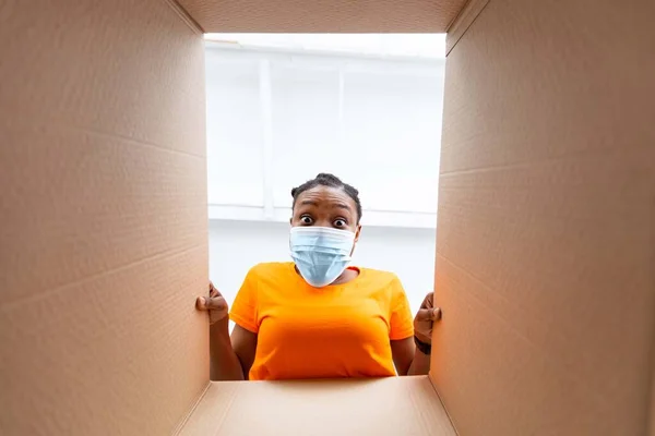Online winkelen en levering tijdens coronavirus quarantaine. Vrolijke Afro vrouw in masker unboxing kartonnen pakket thuis — Stockfoto