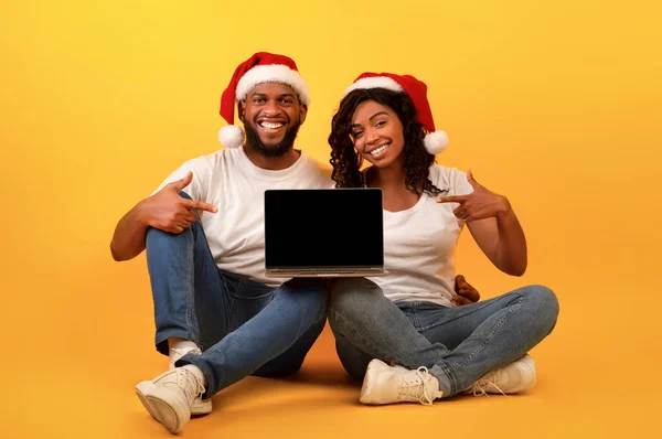 Spännande afrikansk amerikansk par i Santa hattar pekar på laptop med tom skärm, sitter på gul bakgrund — Stockfoto