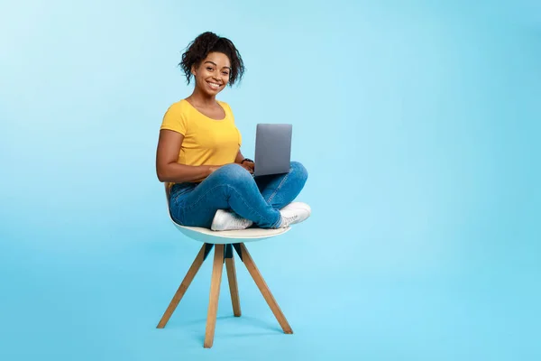 Joyeux jeune Afro dame assise sur la chaise, en utilisant un ordinateur portable, étudier en ligne ou avoir une réunion d'affaires sur fond bleu — Photo