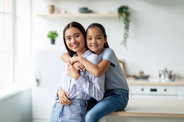 Concepto de ternura y maternidad. Linda chica asiática abrazando a la madre desde la espalda, sentado en la mesa de la cena en la cocina —  Fotos de Stock